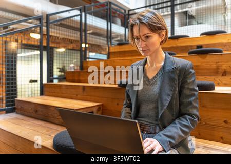 Professional woman in blazer working on laptop on wooden steps in modern coworking space Stock Photo