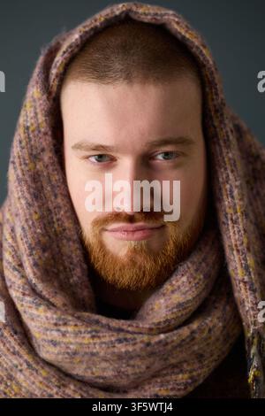 Portrait of young adult Caucasian man with red beard looking at camera while wearing warm scarf on head. Image represents winter fashion and is suitable for apparel marketing, vertical Stock Photo