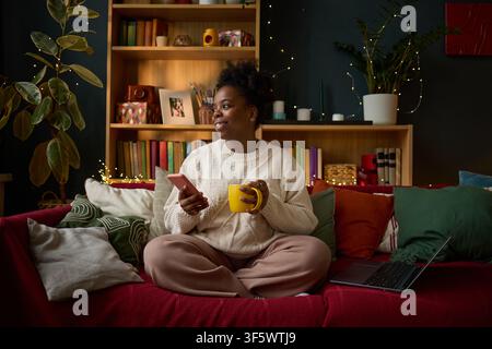 Young adult African American woman sitting on sofa holding smartphone and mug, looking away smiling. Image represents remote work lifestyle, suitable for home office marketing campaigns Stock Photo