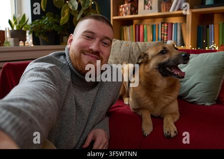Portrait of smiling young adult Caucasian man taking selfie with his dog on sofa at home. Concept of pet ownership and companionship, suitable for veterinary or pet food advertising Stock Photo