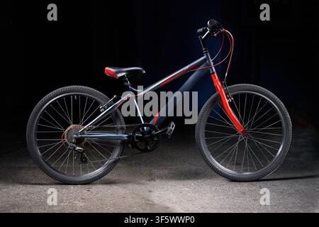 Modern teen mountain bike on concrete ground with dark backdrop. Blue matte steel frame pairs with bright red front fork, classic spoked wheels, knobb Stock Photo