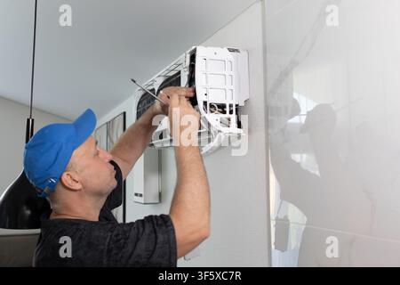 Technician man repairing air conditioning unit on wall. Professional maintenance worker using screwdriver. Climate control system service at home. Daytime soft lighting side view shot. Stock Photo