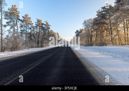 A black asphalt winter road in the middle of the forest, an empty winter road goes into the distance between snow-covered trees at dawn Stock Photo
