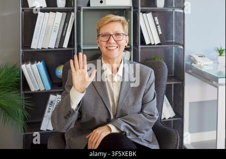 Smiling business professional waving hello during video call in modern office workspace Stock Photo