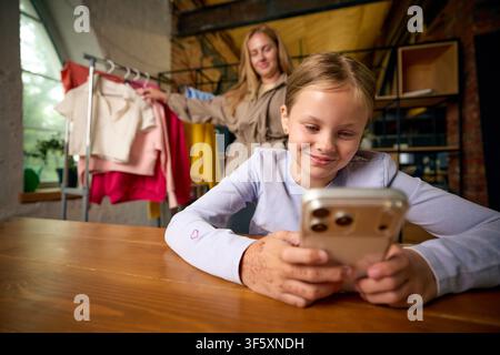 Girl focused on smartphone while mother shows clothing at home Stock Photo