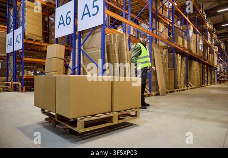 Warehouse worker moving cardboard boxes on pallet trolley in storage aisle Stock Photo