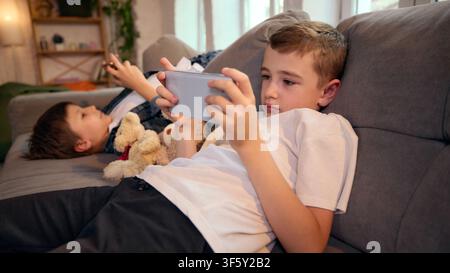 School-age boy using smartphone while sibling rests beside him on sofa Stock Photo