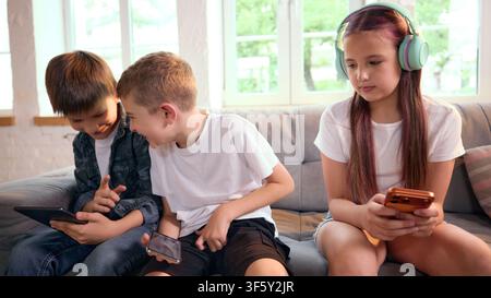Three school-age friends using smartphones together while sitting on sofa Stock Photo