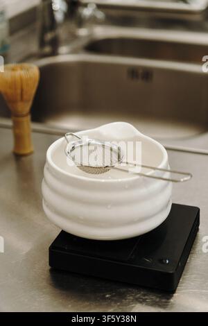 Fine mesh tea strainer resting on a ceramic bowl placed on a digital kitchen scale. Coffee and tea preparation tools arranged on a stainless steel Stock Photo