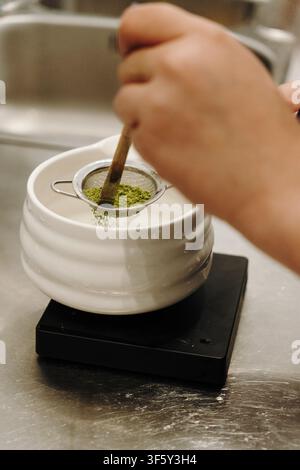 Bamboo whisk gently presses matcha powder through a fine mesh strainer into a ceramic bowl. Traditional tea preparation captured in close-up. Stock Photo