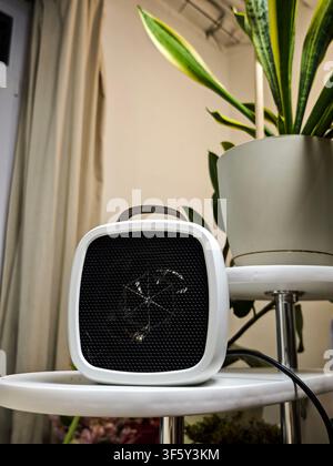 A portable electric space heater sits on a small table beside a tall plant, ready to provide heat in the indoor space during winter. Stock Photo