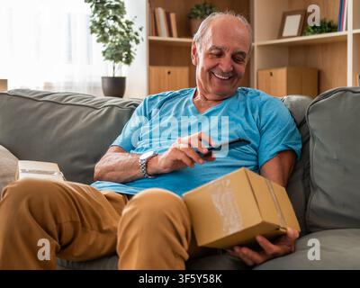 Happy senior man sitting on sofa and scanning QR code on delivered cardboard box with smartphone. He is confirming online order arrival at his home. Stock Photo