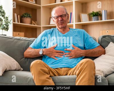 Senior man sitting on sofa in his living room and holding his abdomen with a painful expression. Stock Photo