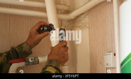 worker adjusts iron heating pipes with a hammer. A light switch is visible on the wall. During the renovation, a plumber, wearing gloves Stock Photo