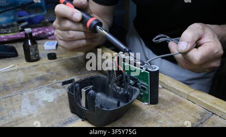 mechanic repairs a battery pack in a workshop. He holds a soldering iron, carefully soldering contacts to the electronic circuit board with wires Stock Photo