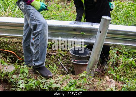 A construction worker installing metal guardrail along roadside with tools and buckets in grassy area, protective gloves and workwear visible Stock Photo