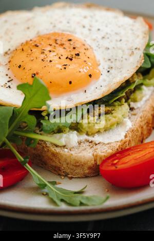 Close up of avocado toast layered with avocado, cheese, arugula and a fried egg. Fresh vegetables complete a nutritious breakfast. Stock Photo