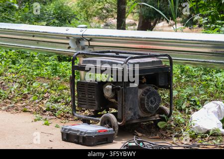 A portable generator on grassy roadside with repair toolbox and cables, rugged metal frame and wheels, outdoor construction equipment providing emerge Stock Photo