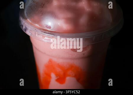 A strawberry milkshake with whipped foam and syrup drizzle in plastic cup under low light, creamy texture and glossy dome lid Stock Photo