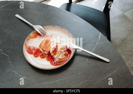 Top view of partially eaten cottage cheese pancakes with berry sauce on a ceramic plate in a modern cafe. Real dining moment showing leftovers after Stock Photo