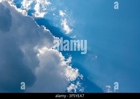 Bright sun rays stream through large white clouds in a clear blue sky. Stock Photo