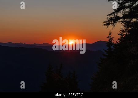 A stunning sunset view over a mountain range. The sun, partially obscured by clouds, casts a warm orange glow across the sky and illuminates the peaks Stock Photo