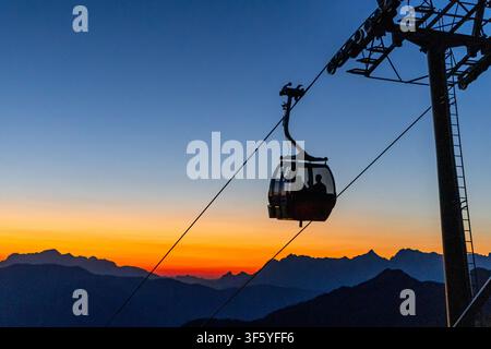 A silhouette of a gondola car ascends against a vibrant orange and blue sunset sky. The cable car is suspended between two towering pylons, with the r Stock Photo