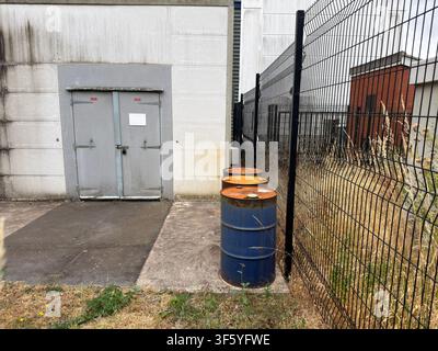 Three weathered industrial steel drums beside security fencing and warehouse loading doors in an anonymous British industrial estate. Stock Photo