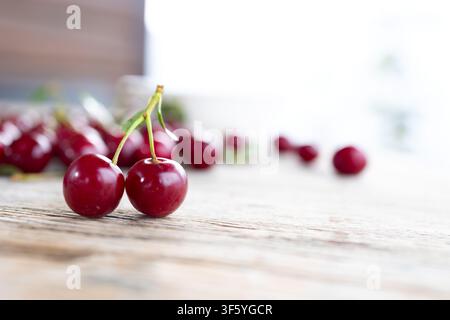 Pair of glossy ripe cherries with green stems positioned on weathered wooden table with scattered out of focus cherries in soft bright background, natural daylight highlights deep red fruit skin Stock Photo