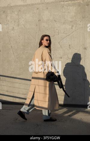 Stylish woman in a long beige trench coat and jeans walking past a textured concrete wall during golden hour. Dramatic, long shadows. Outdoor, full le Stock Photo