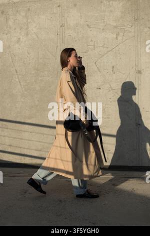Profile view of a stylish woman in a long beige trench coat and jeans adjusting her sunglasses as she walks past a concrete wall. Warm sunset light wi Stock Photo