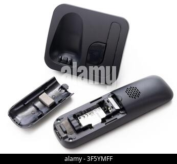 A disassembled black cordless phone with its battery cover removed lies next to its charging base on a white surface Stock Photo