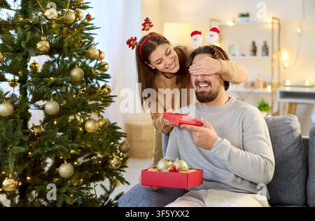 Christmas exchange of gifts in couple, wife covering eyes of husband opening box Stock Photo