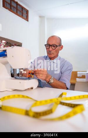 Mature man working diligently with a sewing machine, carefully cutting fabric with scissors on a white table with a measuring tape Stock Photo