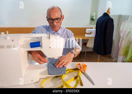 Mature man focused on sewing a denim fabric piece using a sewing machine, surrounded by tailoring tools and finished garments Stock Photo