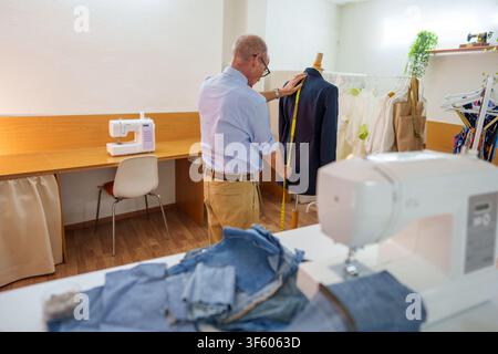 Mature tailor measuring jacket on mannequin, crafting custom garment in workshop, focused on precision and craftsmanship Stock Photo