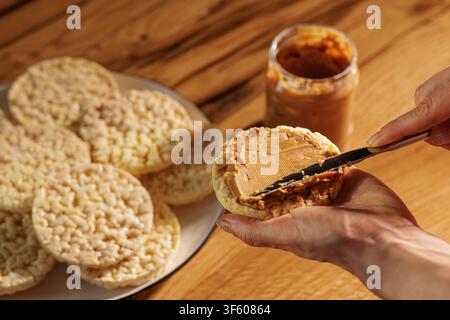 Close view of a hand spreading creamy peanut butter onto a rice cake with a knife, with additional plain cakes and open jar in warm kitchen light. Stock Photo