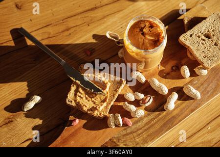 Jar of peanut butter with knife spreading paste on crunchy toast surrounded by whole peanuts on a wooden table in warm natural light. Stock Photo