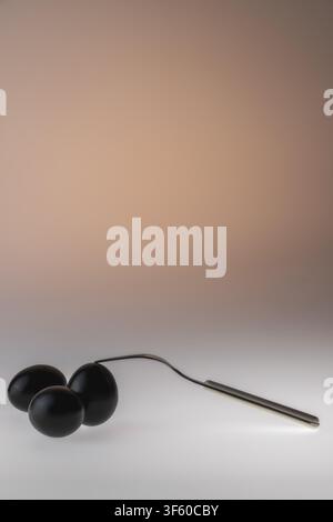 Vertical studio still life of two black painted eggs resting on a curved metal spoon against a warm peach gradient background. The paired dark eggs on Stock Photo
