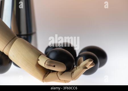 Close-up studio still life of a wooden artist mannequin hand holding black painted eggs with a stainless steel moka pot visible in the background on a Stock Photo