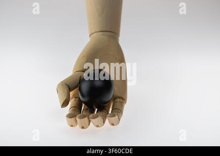 Vertical studio still life of a wooden artist mannequin hand with fingertips pinching a single black painted egg from below against a neutral backgrou Stock Photo