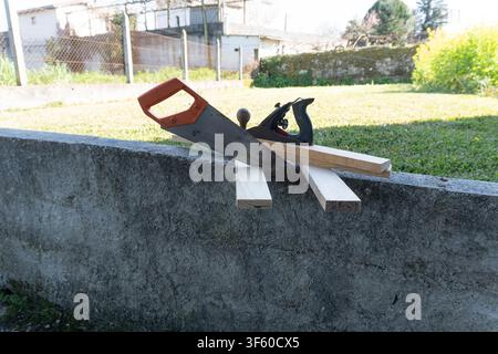 Hand saw and wood plane resting on timber boards across concrete wall in sunny backyard garden Stock Photo