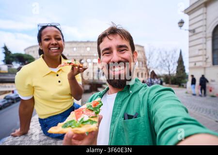 Happy couple taking a fun selfie while eating pizza in front of colosseum in Rome. Stock Photo