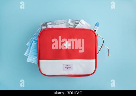 Red and white first aid bag filled with pills, masks, and thermometer. Stethoscope partially visible. Clean, bright lighting, flat lay composition. Es Stock Photo