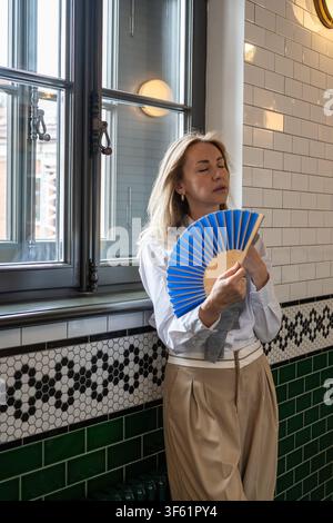 Woman feeling suffering from heatstroke and dizziness indoors, waving paper fan in hand to cool down Stock Photo