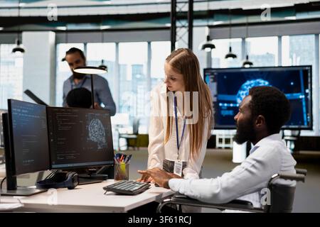 Upbeat technician with disability and junior dev in inclusive office automating tasks with AI scripts. Worker in wheelchair and young intern using artificial intelligence to do feature implementation Stock Photo