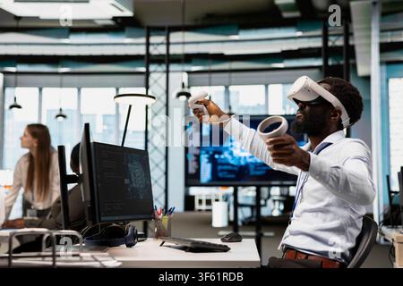 Developer wearing VR goggles at office desk developing programs using AI automation. Tech company worker using virtual reality equipment, debugging applications with artificial intelligence Stock Photo