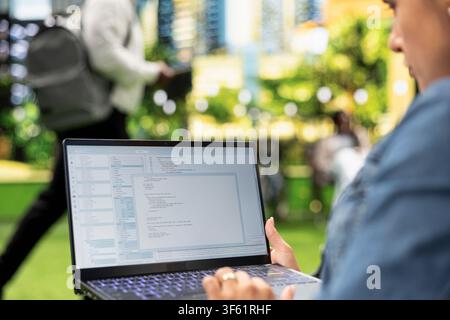 Close up of person working with programming interface and source code on laptop, representing software development and flexible IT work in city park. Over-the-shoulder view. Stock Photo