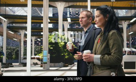 Work colleagues talking and walking inside modern bus terminal, enjoying coffee beverages. Business travel companions having conversation in coach station and holding takeaway drink cups Stock Photo