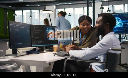 Cheerful manager and employee with paraplegia in inclusive workplace developing software applications. African american technician and colleague with disability using AI to solve bottlenecks, camera A Stock Photo